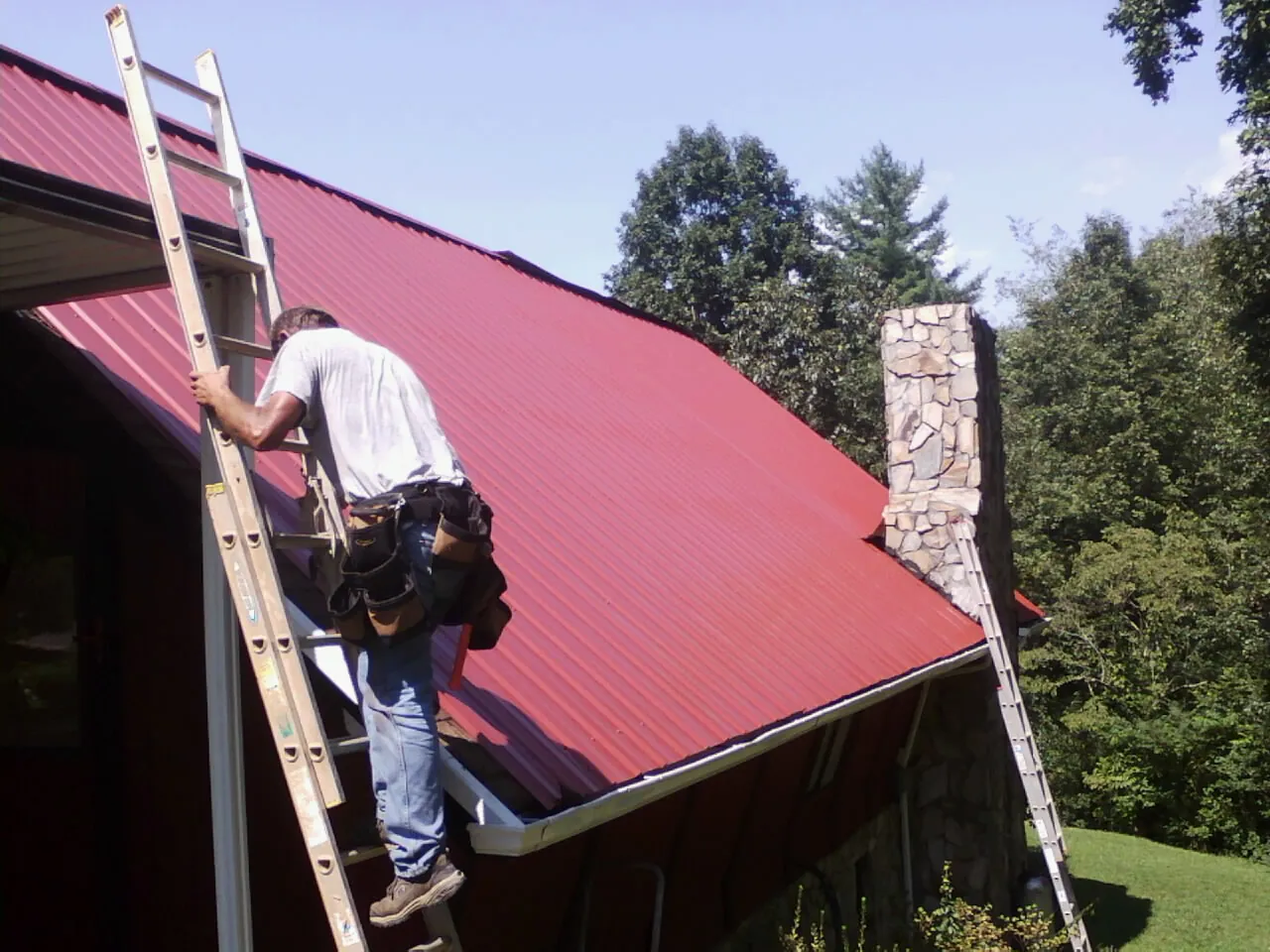 Roofer on a ladder beside a red metal roof and stone chimney — Soffit & Fascia Repair by Roofing Brothers in Coaldale
